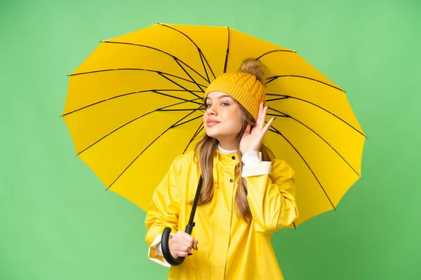 若いです女の子とともにレインプルーフコートと傘上の隔離されたクロマキー背景聞きます何かによって手を置くことによって耳 — ストック写真