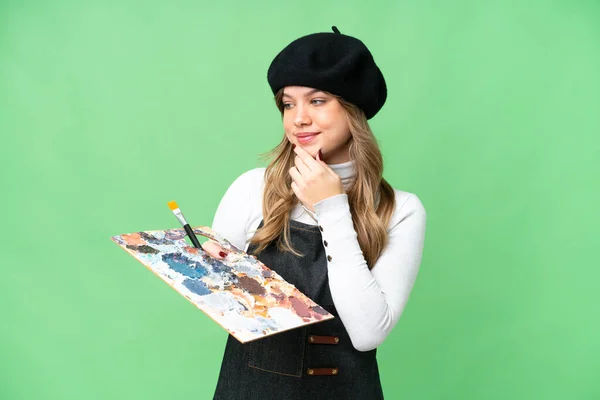年轻的艺术家女孩拿着调色板 在孤立的彩色关键背景上看着侧面 微笑着 — 图库照片