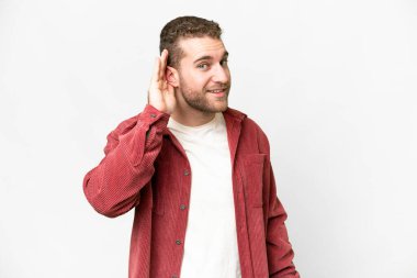 Genç, sarışın, soyutlanmış beyaz arka planda bir şey dinlerken elini kulağına koyuyor.