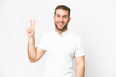 Genç yakışıklı sarışın adam izole edilmiş beyaz arka planda gülümsüyor ve zafer işareti gösteriyor.