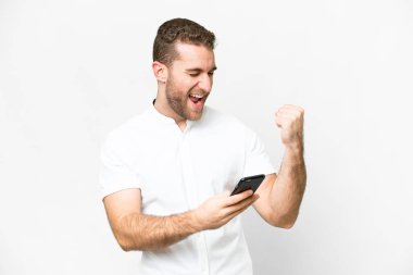 Genç yakışıklı sarışın adam izole edilmiş beyaz arka planda ve telefon zafer pozisyonunda.