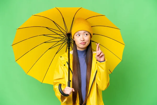 Young Asian woman with rainproof coat and umbrella over isolated chroma key background with fingers crossing and wishing the best