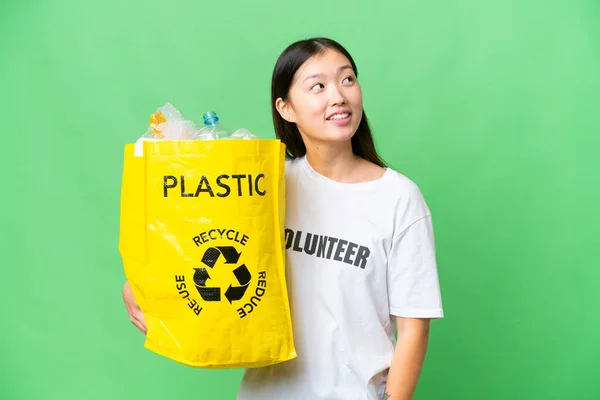 Young Asian woman holding a bag full of plastic bottles to recycle over isolated chroma key background looking up while smiling