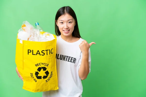 Young Asian woman holding a bag full of plastic bottles to recycle over isolated chroma key background pointing to the side to present a product