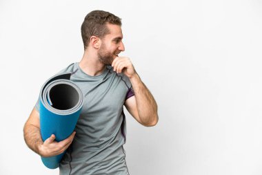 Genç sporcu yoga derslerine giderken elinde paspas tutarak izole edilmiş beyaz arka plan üzerinde bir fikir üretiyor ve bir tarafa bakıyor.