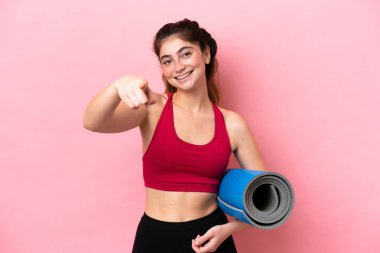 Genç sporcu kadın yoga derslerine giderken elinde mutlu bir ifadeyle önü gösteren bir minder tutuyor.