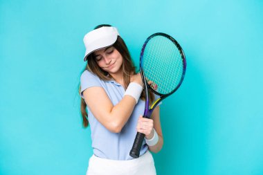 Genç tenisçi kadın mavi arka planda izole edilmiş, omuz ağrısından muzdarip, çabaladığı için.