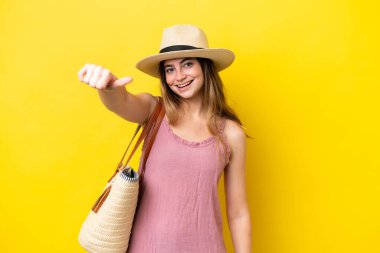 Young caucasian woman holding a beach bag isolated on yellow background giving a thumbs up gesture
