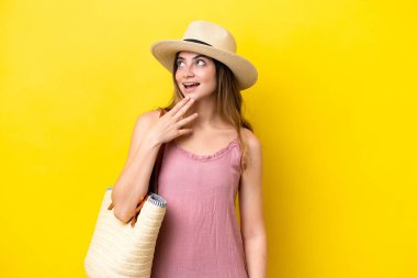 Young caucasian woman holding a beach bag isolated on yellow background looking up while smiling