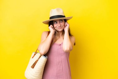 Young caucasian woman holding a beach bag isolated on yellow background frustrated and covering ears