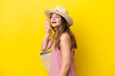 Young caucasian woman holding a beach bag isolated on yellow background listening to something by putting hand on the ear