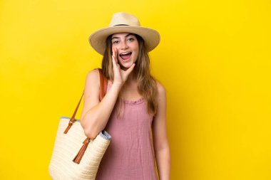 Young caucasian woman holding a beach bag isolated on yellow background shouting with mouth wide open