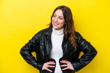 Sarı arka planda izole edilmiş genç beyaz kadın kolları kalçasında ve gülümsüyor.