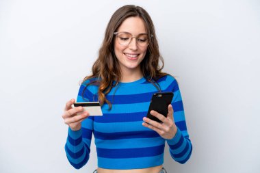Beyaz arka planda izole edilmiş genç beyaz bir kadın cep telefonuyla kredi kartıyla alışveriş yapıyor.