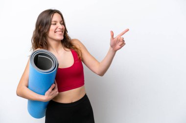 Beyaz arka planda izole edilmiş bir paspası tutarak yoga derslerine giden beyaz, genç bir kadın parmak göstererek bir ürün sunuyor.
