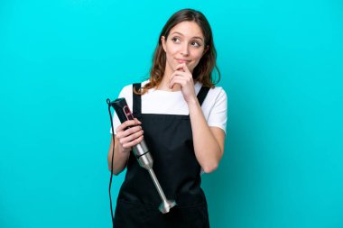 Beyaz tenli genç aşçı kadın el blenderi kullanıyor. Mavi arka planda izole edilmiş ve yukarı bakıyor.