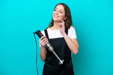 Beyaz tenli genç aşçı kadın el blenderi kullanıyor. Mavi arka planda izole edilmiş, yukarı bakarken bir fikir düşünüyor.