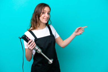 Beyaz tenli genç aşçı kadın el blenderi kullanıyor. Mavi arka planda izole edilmiş. Parmağını yan tarafa gösteriyor.