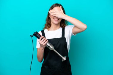 Beyaz tenli genç aşçı kadın el blenderi kullanıyor. Mavi arka planda, gözleri elle kapatıyor. Bir şey görmek istemiyorum.