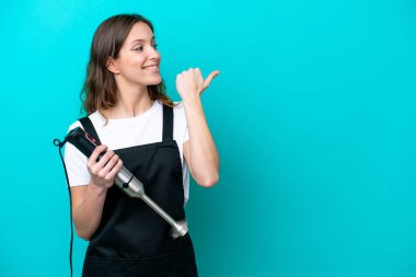 Beyaz tenli genç aşçı kadın mavi arka planda izole edilmiş el blenderi kullanıyor.