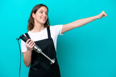 Beyaz tenli genç aşçı kadın mavi arka planda izole edilmiş el blenderi kullanıyor.