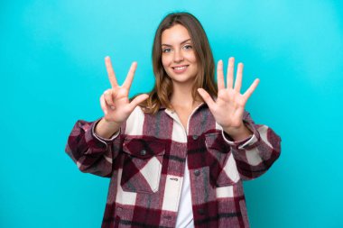 Mavi arka planda izole edilmiş genç beyaz kadın parmaklarıyla sekiz sayıyor.