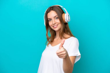 Genç beyaz kadın mavi arka planda izole edilmiş müzik dinliyor ve başparmağını kaldırıyor.