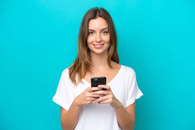 Mavi arka planda izole edilmiş genç beyaz kadın kameraya bakıyor ve cep telefonunu kullanırken gülümsüyor.