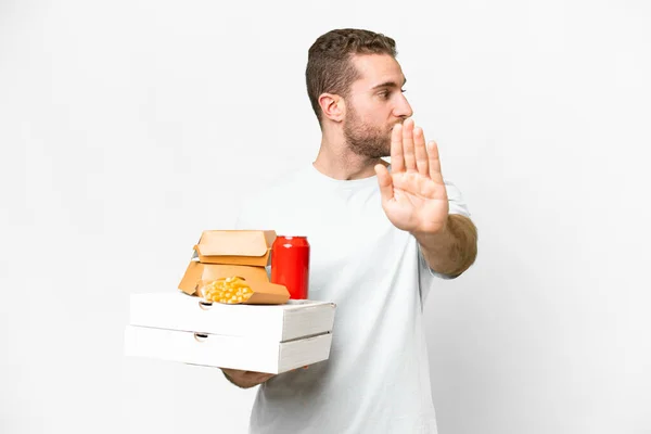 年轻英俊的金发男子举着比萨饼和汉堡包 在孤立的背景下做着停止姿势 感到失望 — 图库照片