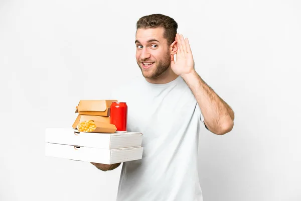 年轻英俊的金发碧眼的男人举着比萨饼和汉堡包 在与世隔绝的背景下听着什么 用手捂住耳朵 — 图库照片