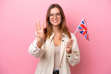 İngiltere bayrağı taşıyan beyaz bir genç kız pembe arka planda izole edilmiş mutlu ve parmaklarıyla üçe kadar sayıyor.