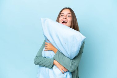 Genç ve güzel bir kadın mavi arka planda pijamalarıyla ve elinde mutlu bir ifadeyle bir yastık tutuyor.
