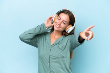 Genç ve güzel bir kadın mavi arka planda pijamalarıyla bir yastık tutuyor ve müzik dinliyor.