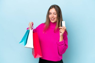Mavi arka planda izole edilmiş genç bir kadın elinde alışveriş çantaları ve bir kredi kartı tutuyor.