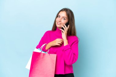 Genç ve güzel bir kadın mavi arka planda izole edilmiş. Elinde alışveriş çantaları var ve cep telefonuyla arkadaşını arıyor.
