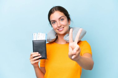 Şişirilebilir seyahat yastığı olan genç ve güzel bir kadın mavi arka planda izole edilmiş gülümsüyor ve zafer işareti gösteriyor.