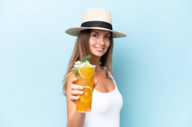 Genç ve güzel bir kadın mavi arka planda mutlu bir ifadeyle izole edilmiş bir kokteyl tutuyor