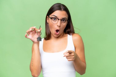 Young caucasian woman holding home keys isolated on green chroma background surprised and pointing front
