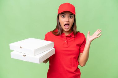 Pizza delivery caucasian woman with work uniform picking up pizza boxes isolated on green chroma background with shocked facial expression