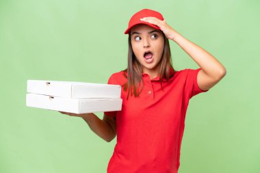 Pizza delivery caucasian woman with work uniform picking up pizza boxes isolated on green chroma background doing surprise gesture while looking to the side