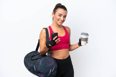 Spor çantalı genç bir kadın beyaz arka planda izole edilmiş. Elinde kahve ve cep telefonu var.