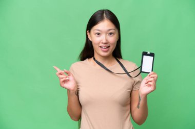 Young Asian woman with ID card over isolated background surprised and pointing finger to the side
