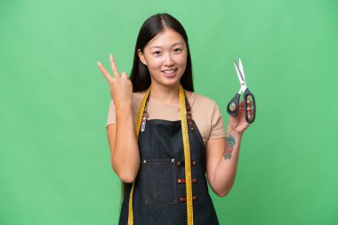 Young Asian seamstress woman over isolated background smiling and showing victory sign
