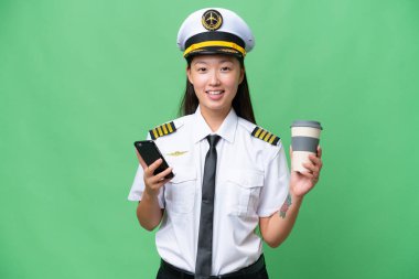 Airplane pilot Asian woman over isolated background holding coffee to take away and a mobile