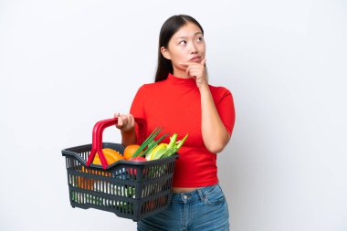 Young Asian woman holding a shopping basket full of food isolated on white background having doubts
