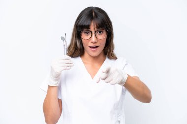 Beyaz arka planda izole edilmiş aletleri tutan beyaz dişçi kadın şaşırmış ve ön tarafı işaret ediyor.