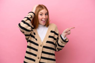 Pembe arka planda izole edilmiş genç, kızıl saçlı kadın şaşırmış ve işaret ediyor.