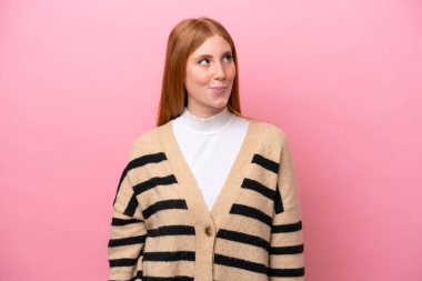 Genç kızıl saçlı kadın pembe arka planda izole edilmiş, yukarı bakarken şüpheleri var.