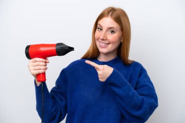 Kızıl saçlı genç bir kadın elinde saç kurutma makinesi tutuyor. Beyaz arka planda izole edilmiş ve onu işaret ediyor.