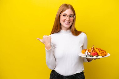 Sarı arka planda izole edilmiş waffle tutan genç kızıl saçlı kadın bir ürünü sunmak için yan tarafı işaret ediyor.